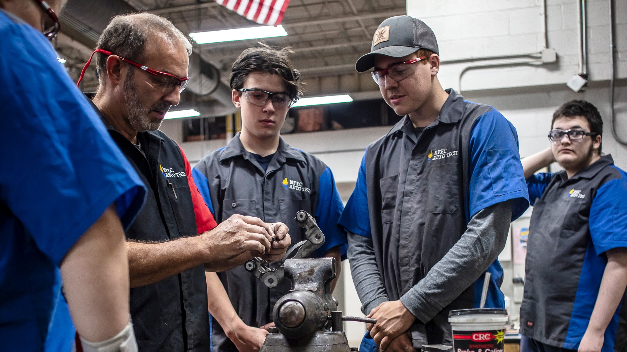 Group of four teen students watching an automotive technician instructor working on a car part