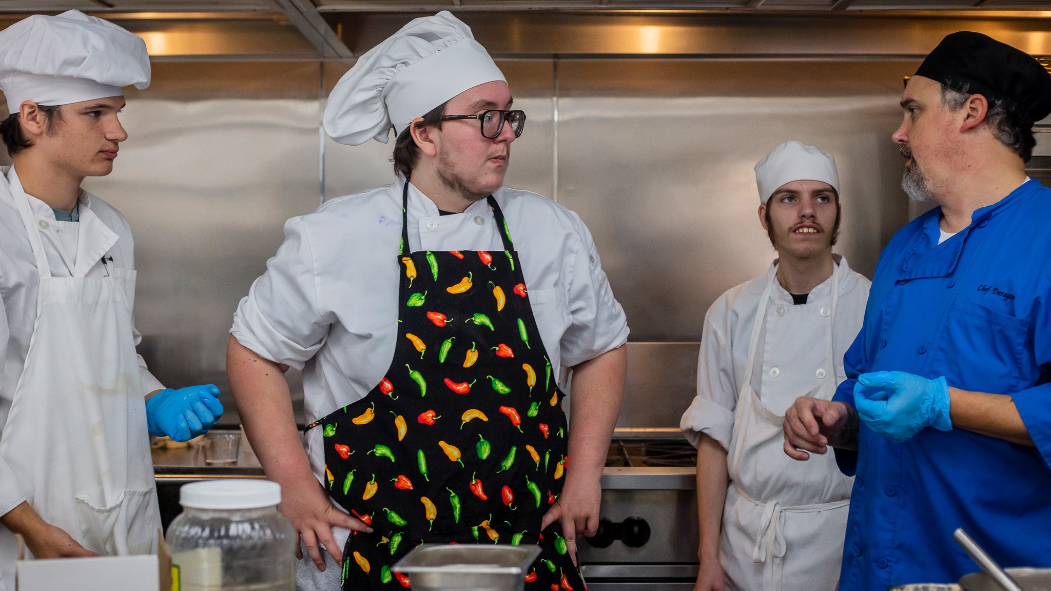 Three teenage culinary students wearing aprons and hats in an industrial kitchen listening to an instructor