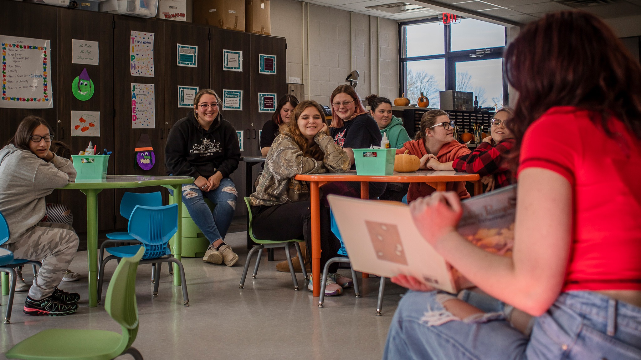 Several teen students sitting at a desk and listening as another reads from a book