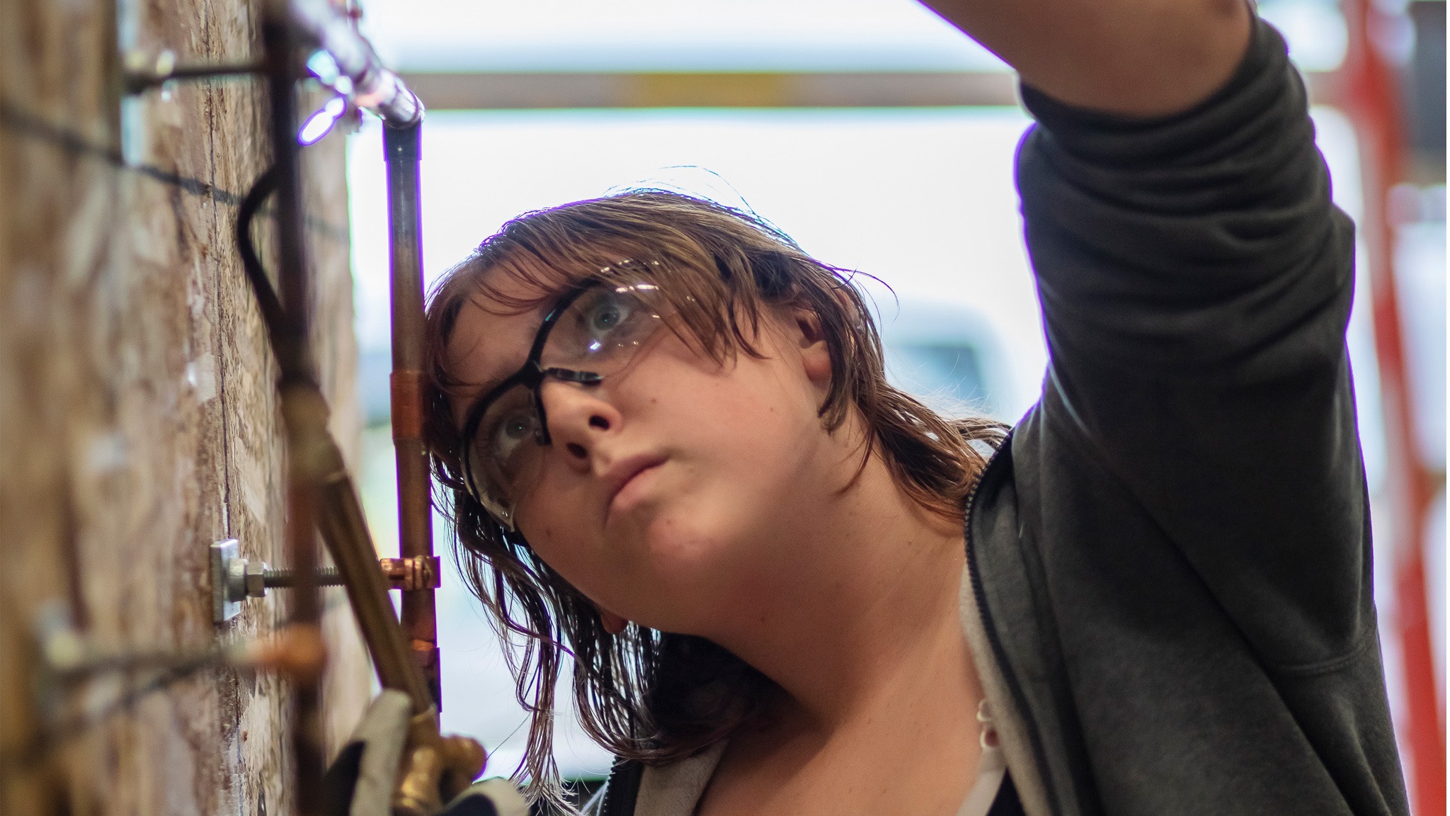 A teen student near copper pipes reaching up to work on a section of pipe