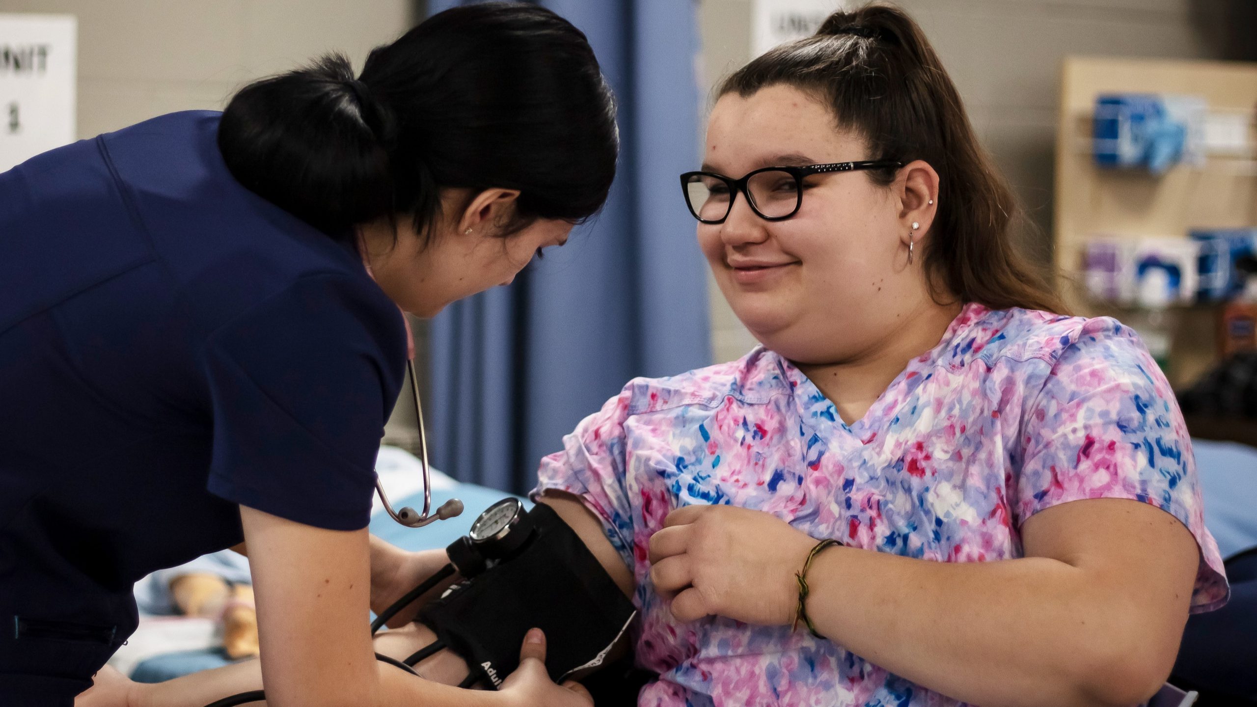 A teen student applying a blood pressure cuff on another student in a training facility room