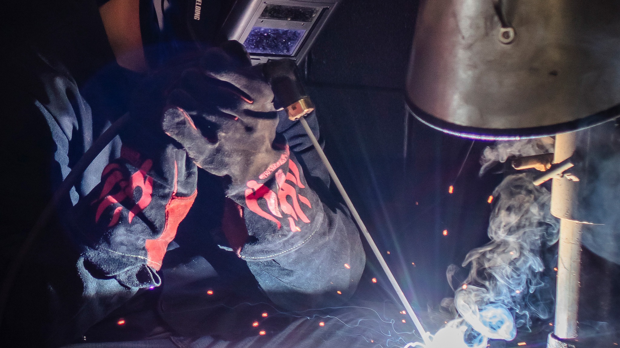 A student wearing a welding helmet welding a piece of metal with smoke rising from the sparks