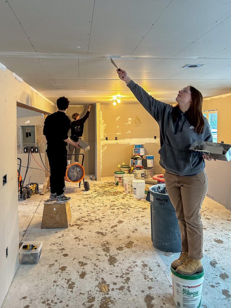 three students working within the modular home, covering up the seems on the ceiling