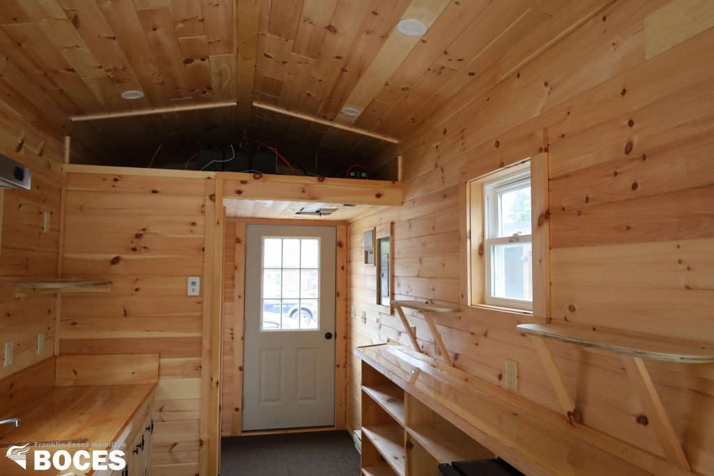 all pine interior of the tiny house, picture shows a small loft area where electrical stuff is, the door to back entrance and where the electrical panel is, the counter top to sink on left of tiny house, right side where the shelfing is, windows on both sides and the toilet near back door.