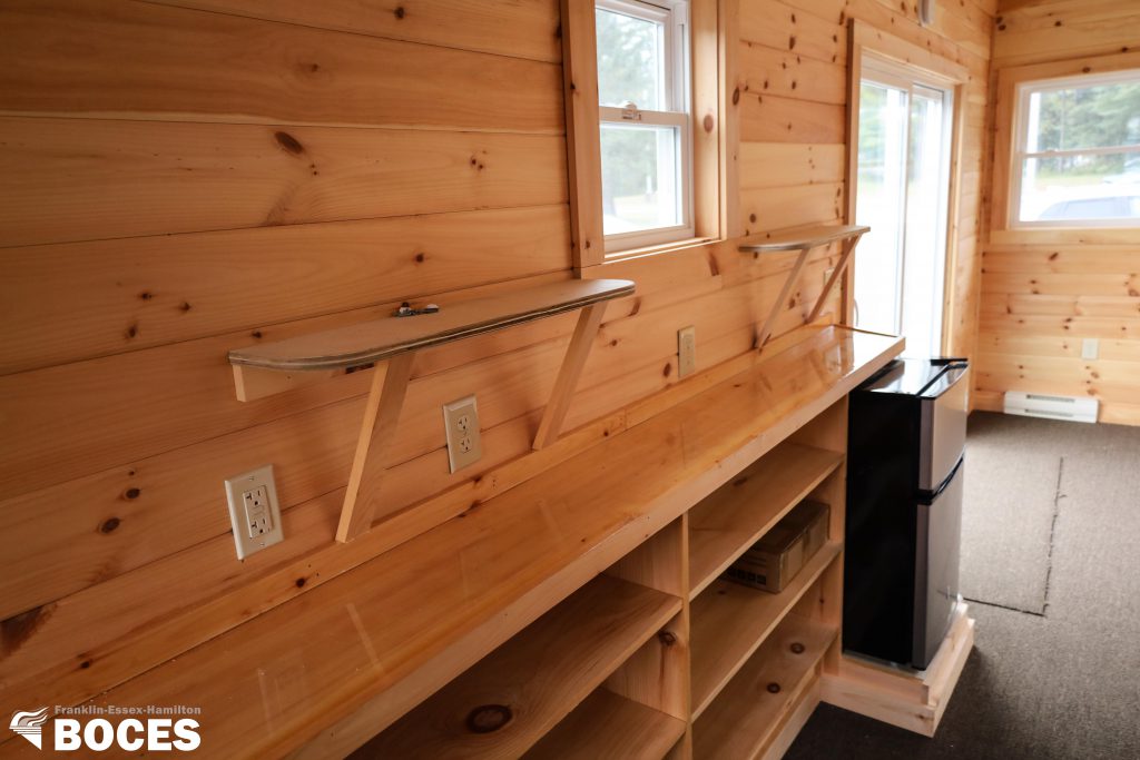 all pine interior of the tiny house, picture shows a mini fridge, shelfing beside the fridge going along the tiny house, some windows, and a sliding door