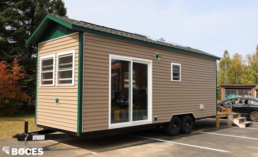 exterior image of the tiny house. shows tan siding, green trim, sliding glass door, a small window and two windows in the front, as well as a hitch that you can use to hook to a truck