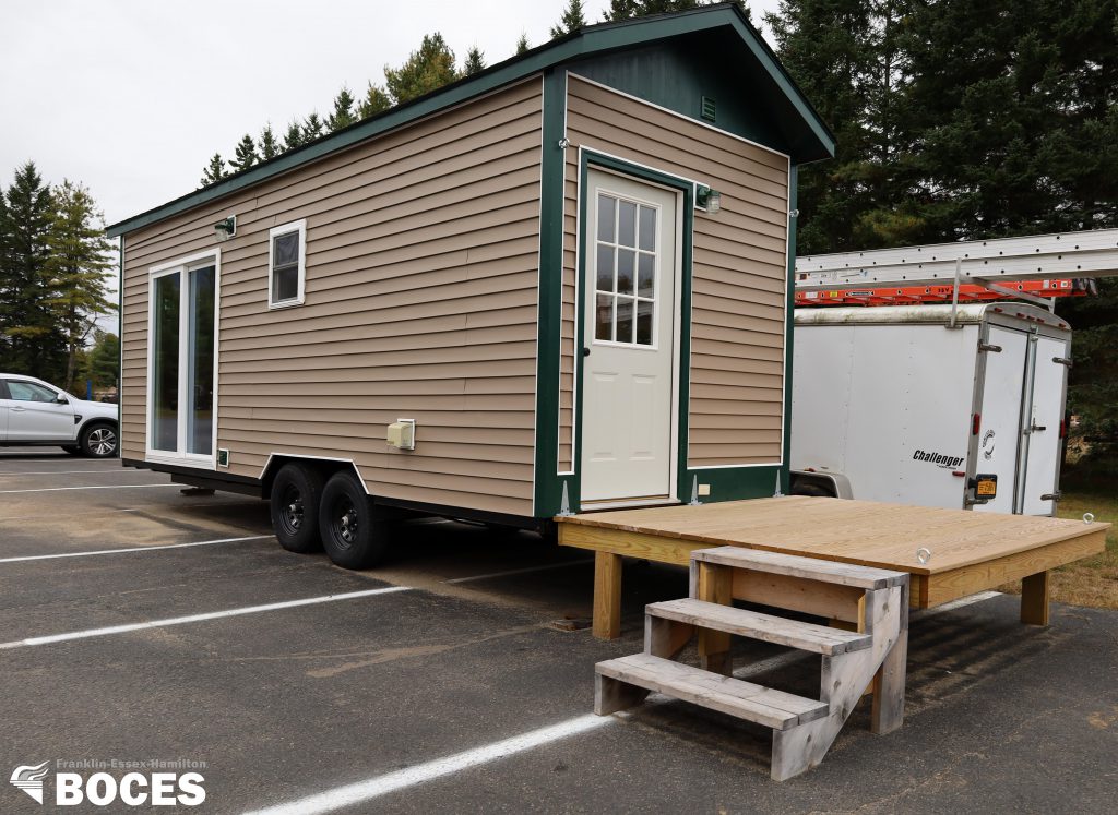 exterior image of the tiny house, it shows a light brown siding, with green trim, mini deck to the back door, a sliding door on the side and a small window
