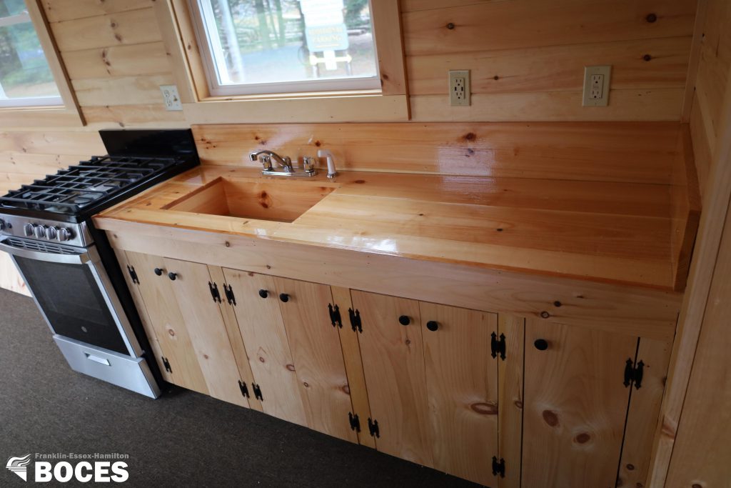 all pine interior of the tiny house, picture shows the counter with cabinets, a sink built in and a gas stove, a window above the sink