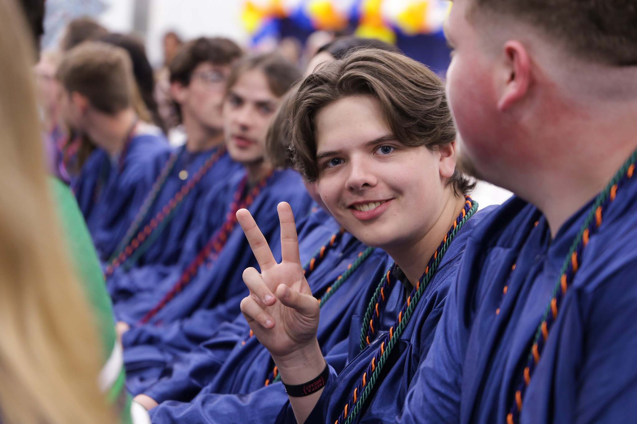 Graduate sitting alongside classmates during ceremony gesturing a peace sign to camera